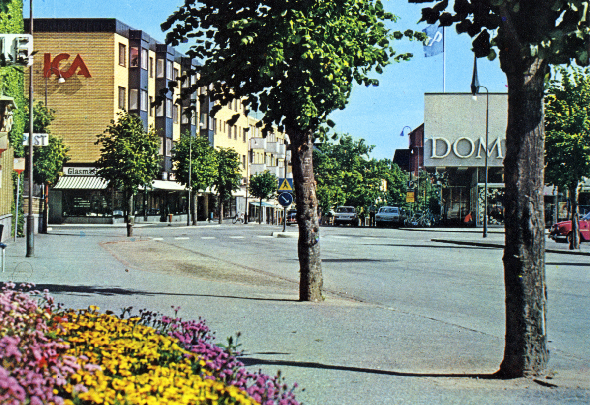 Centralgatan, till vänster det så kallade ICA-huset och till höger Domus. Kvarteren Apotekaren och Plåtslagaren. 

Trafik, sommar.