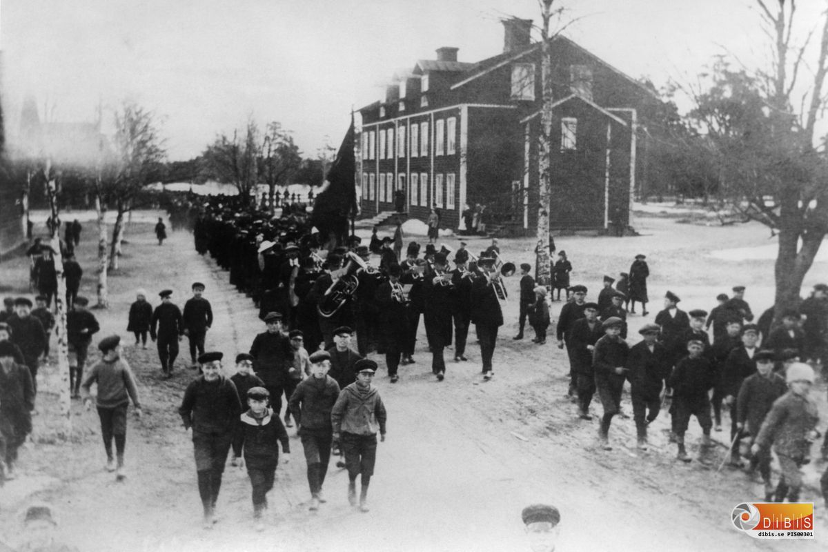 Demonstration 1 maj i Sandarne c:a 1900-1910.
Denna bild var införd i Söderhamns Kuriren den 30/4 1964. Bildtext: Så här såg det ut 1 maj i den s k gamla goda tiden. Bilden har lämnats till Kuriren av Erik Brusven, Sandarne, som själv är en av skolmösseförsedda pojkarna som trippar längst t h på bilden. Händelsen torde härröra omkring femtio år tillbaka i tiden och visar demonstrationståget från Sandarne på väg mot Söderhamn. Bilden är tagen vid den gamla bagargrinden mellan Sandarne och Källskär. Det är Sandarne musikkår som går i spetsen och kortet är taget av J A Lindberg.