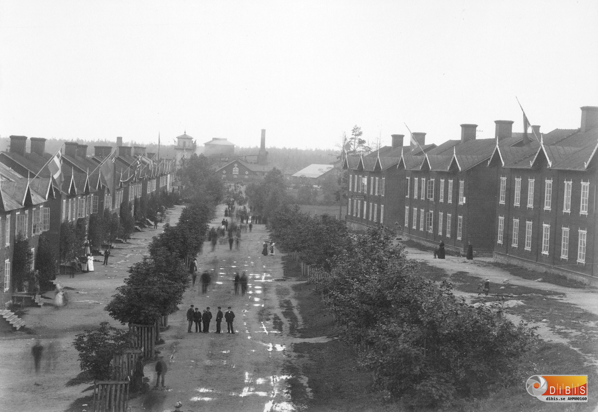 Bostadshus efter Nygatan i Ljusne dekorerade med fasadflaggor inför Kung Oscar II besök, 20 augusti 1890.
I bakgrunden spruthuset och järnverket.