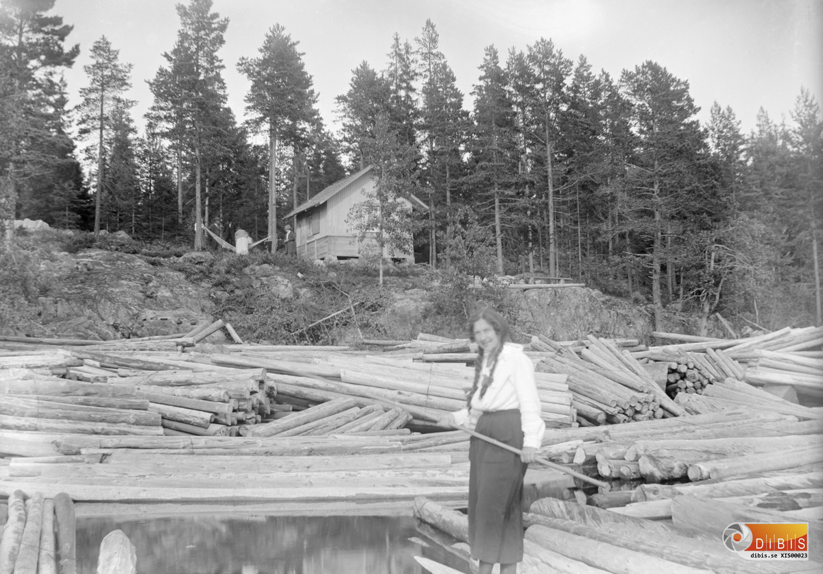 Uppe på berghällen, J Israelssons stuga i Hibo. Timret som har strandat skvallrar om att kortet är taget efter vårfloden 1916 damen är Elisabet Israelsson.