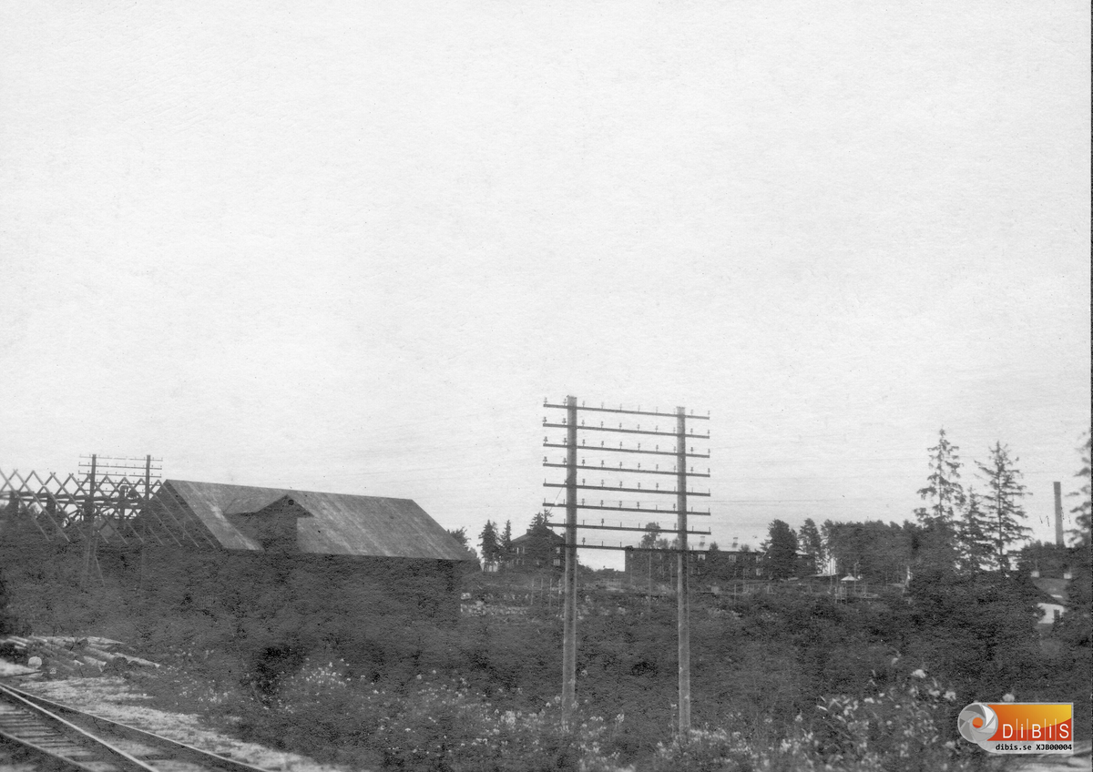 Motiv taget från Kilaforsbanan i augusti 1927. Huset är den så kallade kolladan där träkol lagrades när det var kolmilor på industriområdet, där lagrades även den träkol som kolades ute i skogen av bönder, som sedan körde fram sitt tråkol och fick det inmätt och betalt där.
Bakom telefonstolpen syns Grisbergskasernerna på Bruket där två av dem vid fotograferings tillfället sjunger på sista versen, de rivs och flyttas längst ned på Stenbacka, längst till höger syns sågens skorsten.
