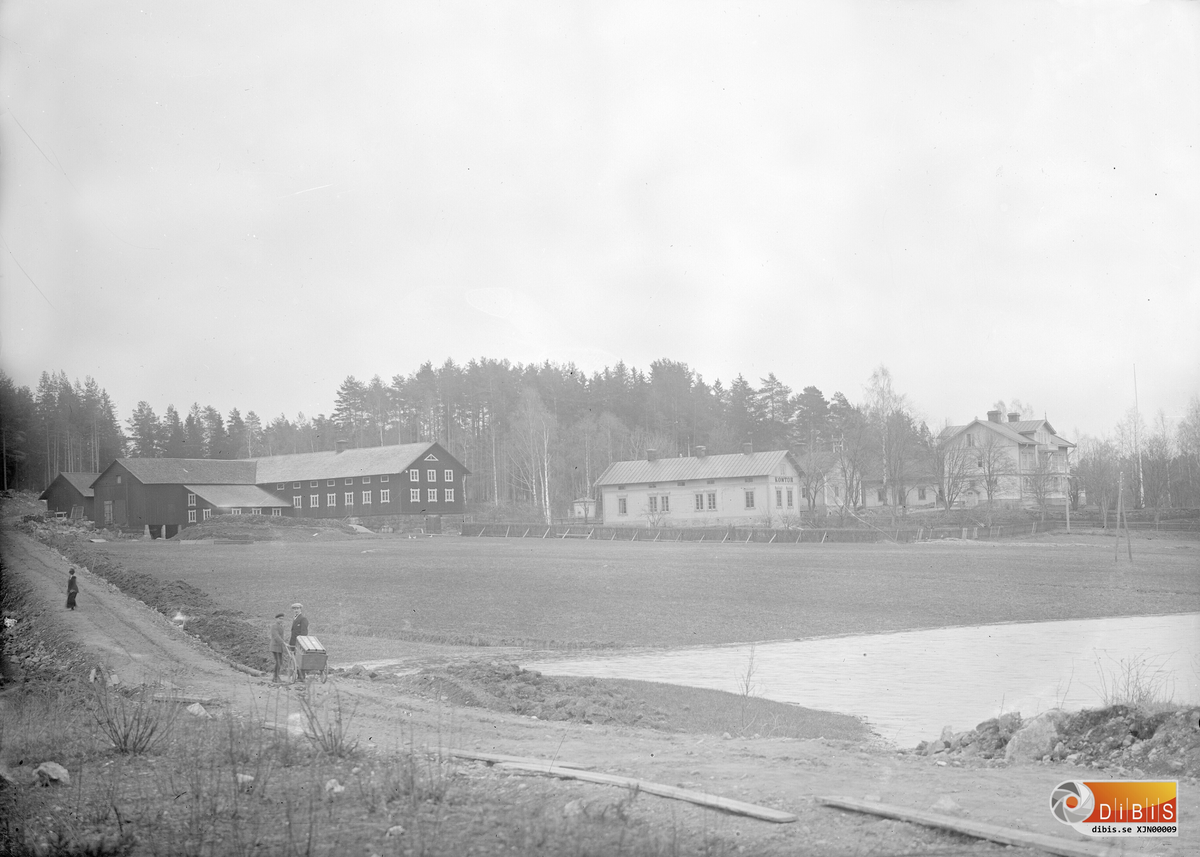 Den nya herrgårdsgatan 1921, med ladugården, sågverkskontoret och herrgården i bakgrunden. Två unga män Gunnar Magnusson f 1896 och Eskil Gudmundsson f 1903 drar på en kärra med en Amerikakoffert som de hämtat på stationen, själv avled emigranten på väg hem med tåg från Göteborg han hette Albin Magnusson f 21/1 1893 emigrerade till USA 1910 och avled 24/11 1921.