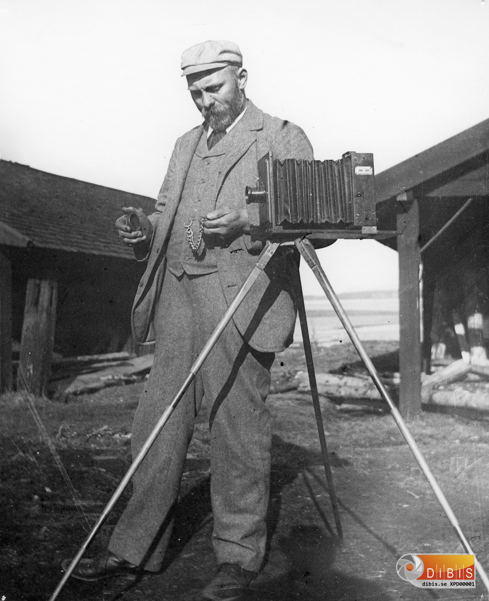 Per Lind, född 26/8 1862, död 25/5 1914. Son till Soldat Jonas Lind och hans hustru Margta Andersdotter, i Östra Höle, Rengsjö. Per Lind var till yrket Kakelugnsmakare, men som ett andra yrke hade han fotografering.  Han arbetade på många platser utanför socknen som kakelugnsmakare och samtidigt fotograferade han, vilket framgår av hans efterlämnade samling av glasplåtar. Per Lind var gift med Mathilda Johansdotter och de hade fyra barn, döttrarna Jenny, Märta, Maria samt sonen Herman. Märta och Maria blev också fotografer, och en del av glasplåtarna har dom tagit. Då det ej går att avgöra vem som har tagit en del av bilderna går alla bilder under beteckningen "Per Linds samling". På denna bild står PER LIND vid sin kamera, platsen obekant. Kameran och stativet, liksom hela glasplåtssamlingen har donerats till Rengsjö Hembygdsförening.
