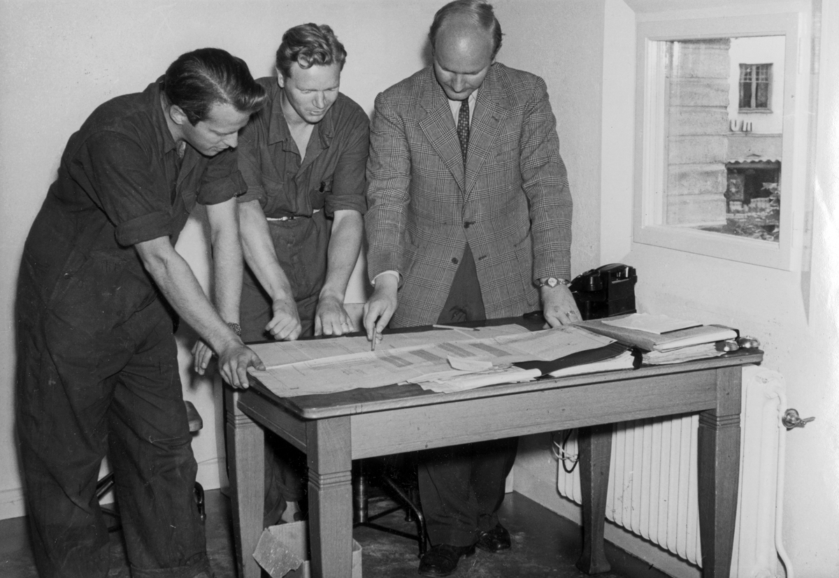 Byggnation av Post- och telehuset, Centralgatan 16, kvarteret Grankotten. Tre män står vid ett skrivbord och tittar på ritningar. Post- och telehuset byggdes 1950-1951 i hörnet Centralgatan - Lövlundsvägen, kvarteret Grankotten. Det ritades av arkitekt Yngve Ahlbom och stod färdigt 1952. 

Yrke, arbete.

Personer på bilden:
Montageledare ingenjör Nyström