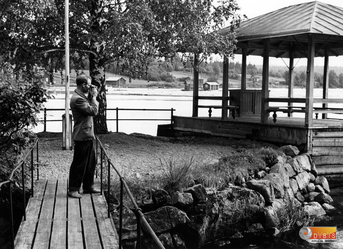 Bilden visar en byggnad som bara kallades Paviljongen. Den låg uppströms ett par hundra meter från nuvarande kraftverk i Bergvik. Om bilden skulle vara fullständig borde även kägelbanan vara med. Kägelbanan och paviljongen användes ofta samtidigt. Kägelspelet avslutades med servering under paviljongens tak eller så var det en plats att vänta på mellan spelen.
Som så mycket annat från Bergviks historia försvann paviljongen när kraftverket byggdes.

När bolagets huvudkontor 1917 flyttades till Söderhamn användes kägelbanan mindre ofta och områdets underhåll blev sämre. Fönsterrutorna blev småpojkarnas eftertraktade byte och i slutet av 1950-talet revs paviljongen. Anläggningen i sin helhet schaktades bort 1960 i samband med kraftverksbygget. Idag är området täckt av en stor kulle av schaktmassor, som likt en forntida vikingagrav rymmer delar av en gången tids kultur.

Borta är även det mesta av vad som syns i bakgrunden. Exempelvis de två byygnaderna på vattnet synliga till vänster om paviljongens bordsliknande möbel.
Den större byggnaden inrymde ett spel med
kraft nog att dra fram virket när motvind störde tappningen genom forsen. Virket drogs i länsar från skiljet i Segersta och tappades i en jämn ström för att i lagom takt nå skiljet i Sunnanå.
Till arbetsplatsen hörde också en liten matsal där bl a Ferdinand Strid tog sina pauser under arbetet.
Byggnaderna på motsatta stranden är alla borta. I dag ligger båtklubbens hamnanläggning ungefär där buskaget tar vid till höger i bilden.
Foto: Sven-Erik Johansson