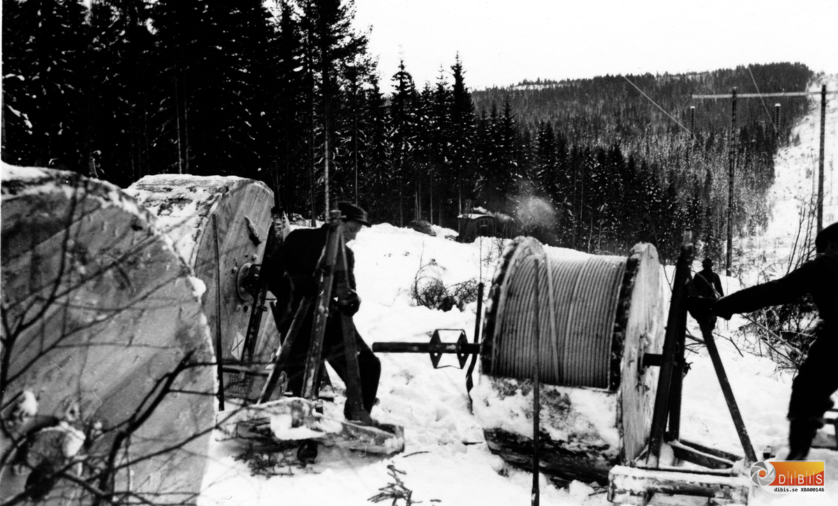 Denna  bild är den första i en rad bilder som skildrar kraftledningsbyggarnas arbete på 1950-talet. Samlingen av bilder kommer från Ingvar Strid, Verkmyra, Bergvik. Han var under många år ansvarig förman för ett antal linjearbetare vid kraftförvaltningen i Bergvik.
Nestorn Lars Wiklund har varit med om granskningen av bilderna. Han var själv linjearbetare och har bistått med sin sakkunskap.
Personen till vänster är sannolikt Olle Wiklund.
I bakgrunden skymtar en rastkoja av den typ som framställdes vid Hurtigs sågverk i Stråtjära. Kojan kunde fällas ihop och transporteras på de medar som fanns under golvet. Tillkomsten av de här funktionella kojorna var revolutionerande för såväl linjearbetare som skogshuggare.
Granskar man originalbilden skymtar också en Volkswagen intill kojan. Den tillhör Ingvar Strid som i början av 1950-talet köpte en sådan bil.