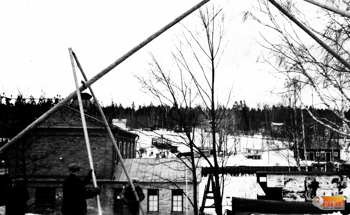 Här pågår arbeten på "kraftens" hemmaplan
d v s lokalerna på norra sidan av älvbron i Bergvik. En kraftledning ska byggas och resning av stolpar pågår. Tegelbyggnaden är ett sedan länge rivet garage som i bottenplanet innehöll en tvättstuga.
Till höger om byggnaderna finns en väl tilltagen anslagstavla. Placeringen kom sig av att vid den här tiden var det gång- och cykelväg till Bergviks station.
Den lilla byggnaden till höger är kallbadhuset, nära till för alla som bodde på Lappstan. Bakom badhuset ligger en av bolagets ekonomibyggnader.
Kiosken tillhörde Sigurd Engelbrektsson. Den skulle senare flyttas till andra sidan av landsvägen.
Brostugan skymtar till vänster. I närheten står en landsvägsbuss som tämligen säkert ingick i Halvar Hasts vagnpark med garage och utgångsläge i Stråtjära.