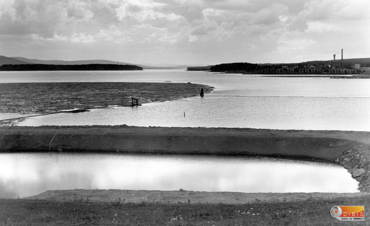 Här har "bassängen" vid .Badkullen färdigställts. Den  byggdes med scaktmassor som kom från bygget av Bergviks kraftverk.
Idén att bygga en badplats kom från Gösta Sundin, då chef för Kraftförvaltningen i Bergvik. I projektet ingick även att uppvärmt vatten, en restprodukt från processen i kraftverket, skulle temperera vattnet i bassängen.
Dessutom skulle sluttningen ner mot bassängen fungera som en plats för solbad.
Med facit i hand kan konstateras att kullen och badet inte fullt ut svarade mot förväntningarna.
Bilden är intressant även ur andra aspekter. I bakgrunden syns fabriken i Vannsätter. Når bilden togs pågick flottningen och fabriken tog hand om råvaran som sorterats ut vid skiljet i Segersta. När veden senare levererades med lastbilar försvann de långa vältorna.
Veden flisades och lagrades bildade ett imponerande berg från vilket en bandtraktor hämtade och levererade råvaran.
Virket i förgrunden skulle senare sorteras vid skiljet i Sunnanå.