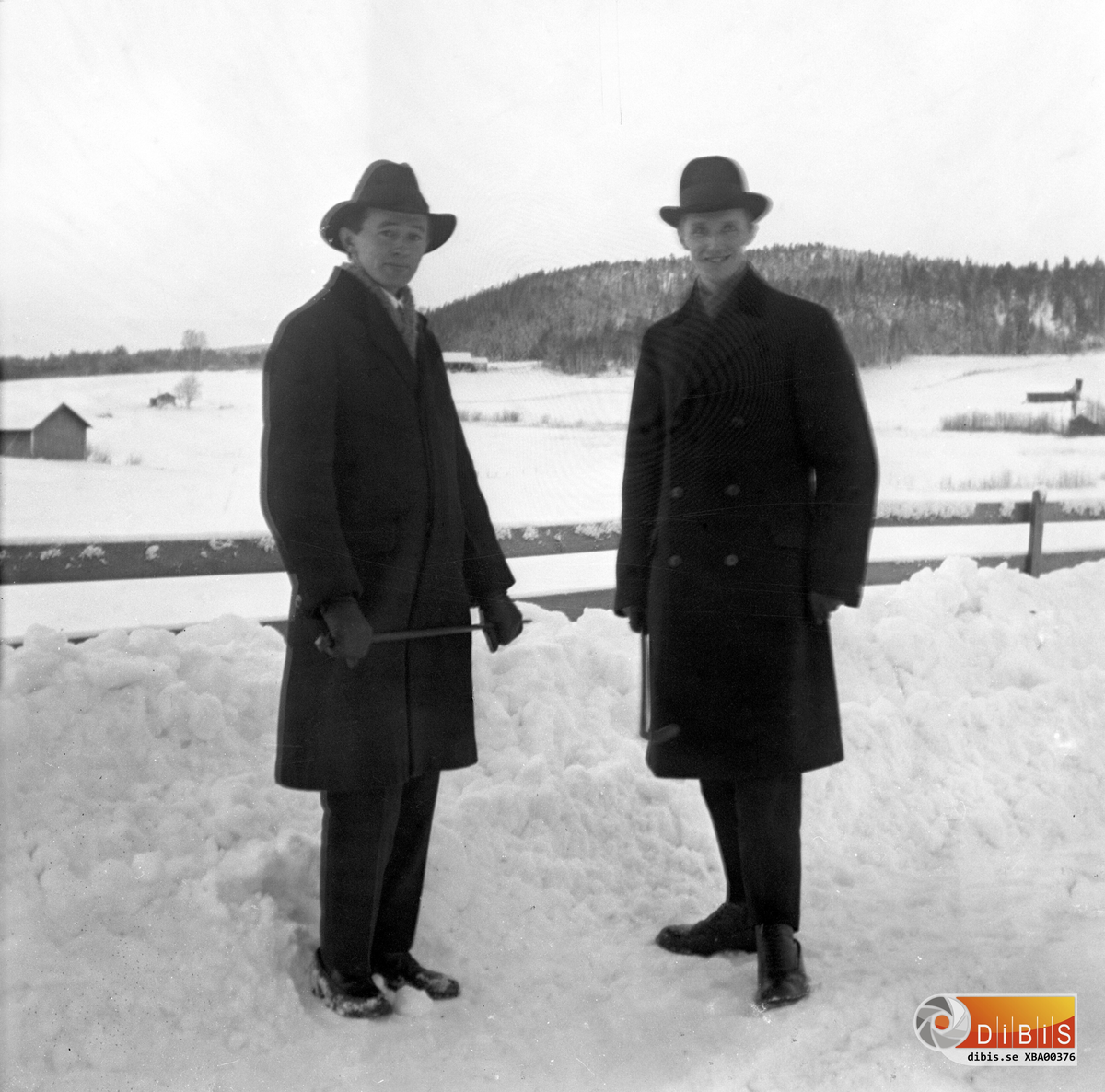 Petrus Zettersten (till vänster) är ute på promenad tillsammans med en god vän. Det är vinter och gott om snö. Bilden avslöjar att plogningen inte var den bästa. Det går också att se några av de lador som fanns på gårdarna i Verkmyra. Byggnaden mot den bakomliggande skogen hör till den gård som då ägdes av Karl Lindberg.