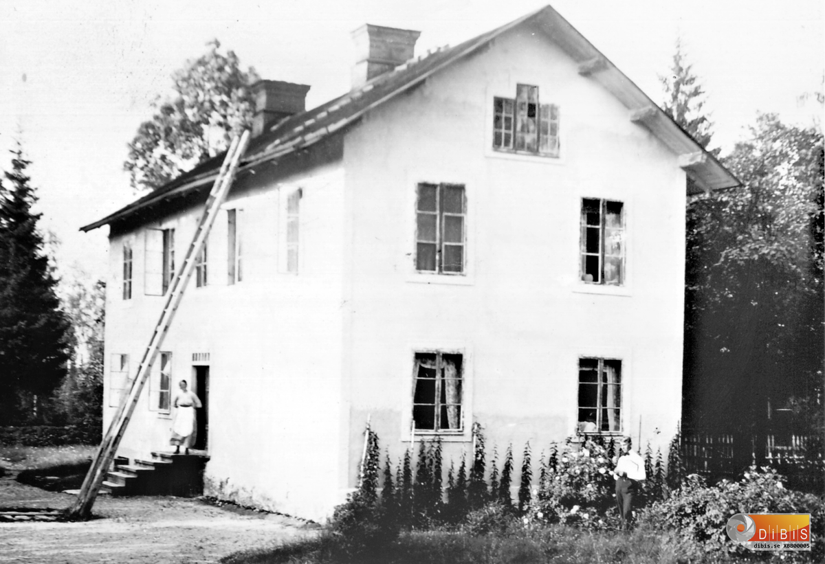 Så såg Karlslunds huvudbyggnad ut från "baksidan" där utbyggnad och veranda idag finns. Man skymtar staketet och vägen till höger. På trappan står Anna Lindblom och den unge mannen bland blommorna är näst äldste sonen Gunnar Lindblom. Bilden är tagen 1918-1919.