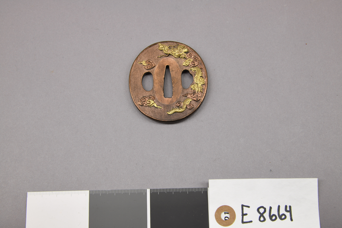 E 8664 Fra protokoll: "Tzuba av rød bronse. Oval. Høy relieff med innlegg i gul kobber. Granulert bund-Nanako. Motiv: flygende drage. Revers: liten drage. Usign. 6,2 x 5,8 cm. Japan."