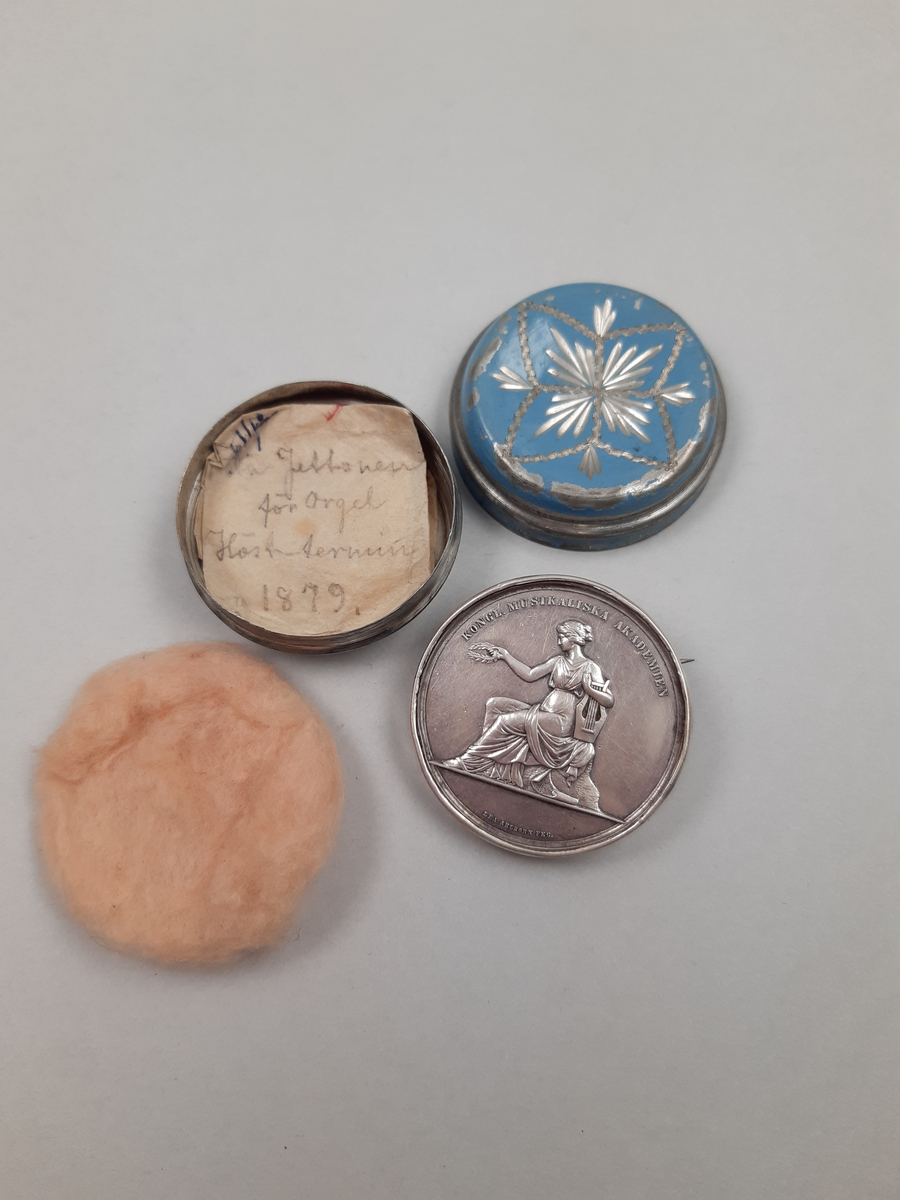 Silvermedalj med relief föreställande musikkonstens musa, omgjord till brosch i ljusblå metallburk med stjärnmönster.
