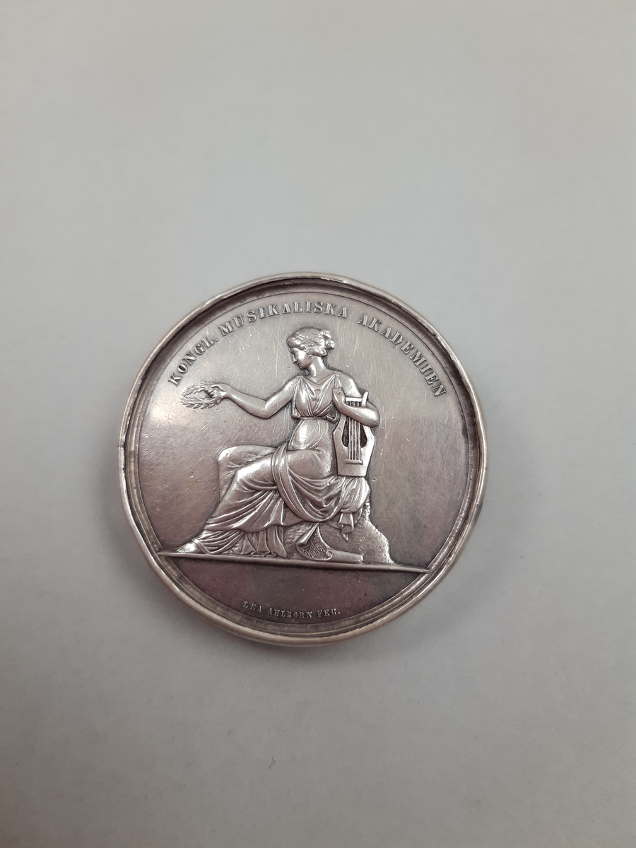 Silvermedalj med relief föreställande musikkonstens musa, omgjord till brosch i ljusblå metallburk med stjärnmönster.
