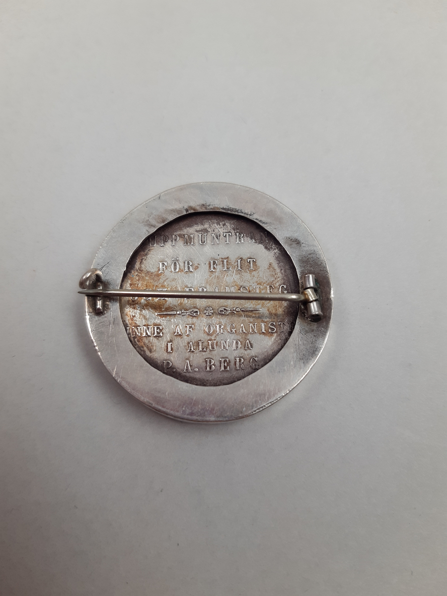 Silvermedalj med relief föreställande musikkonstens musa, omgjord till brosch i ljusblå metallburk med stjärnmönster.
