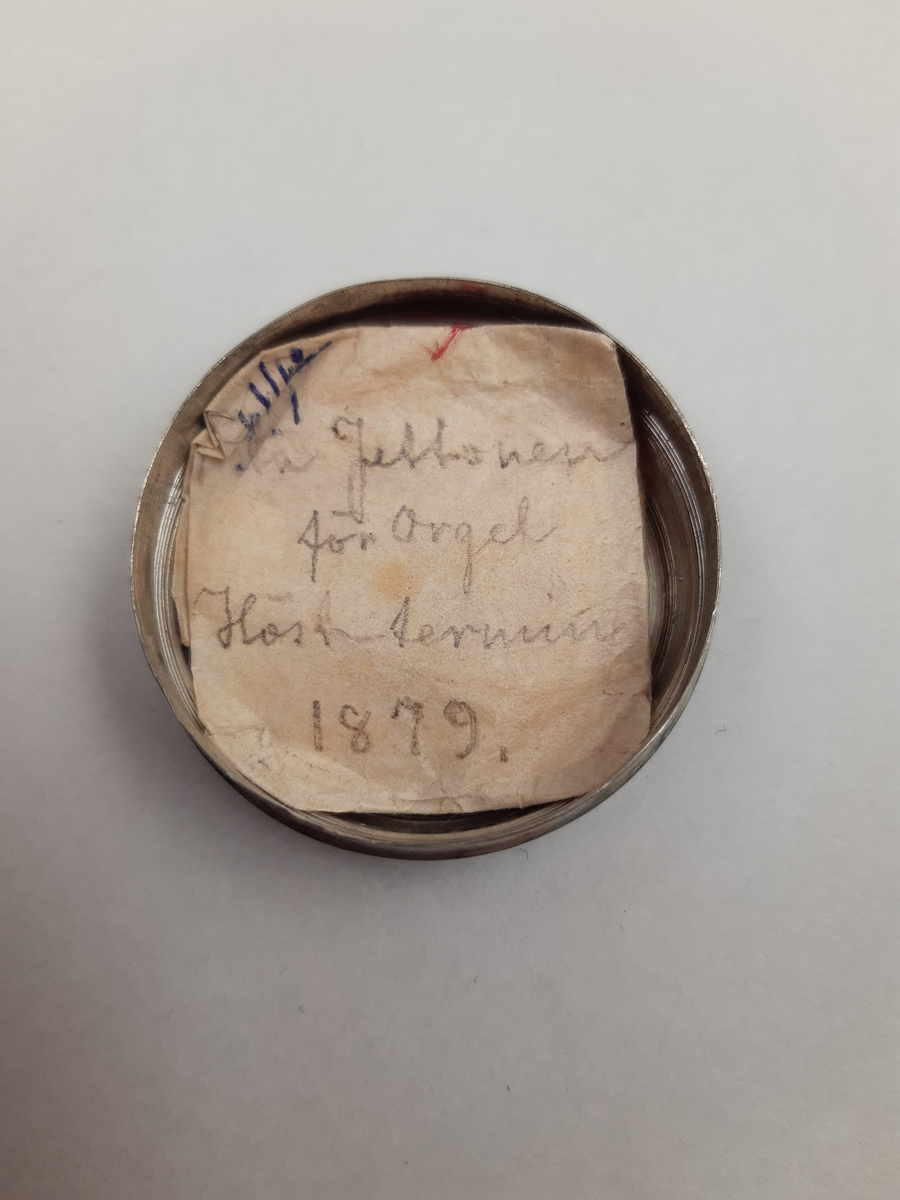 Silvermedalj med relief föreställande musikkonstens musa, omgjord till brosch i ljusblå metallburk med stjärnmönster.
