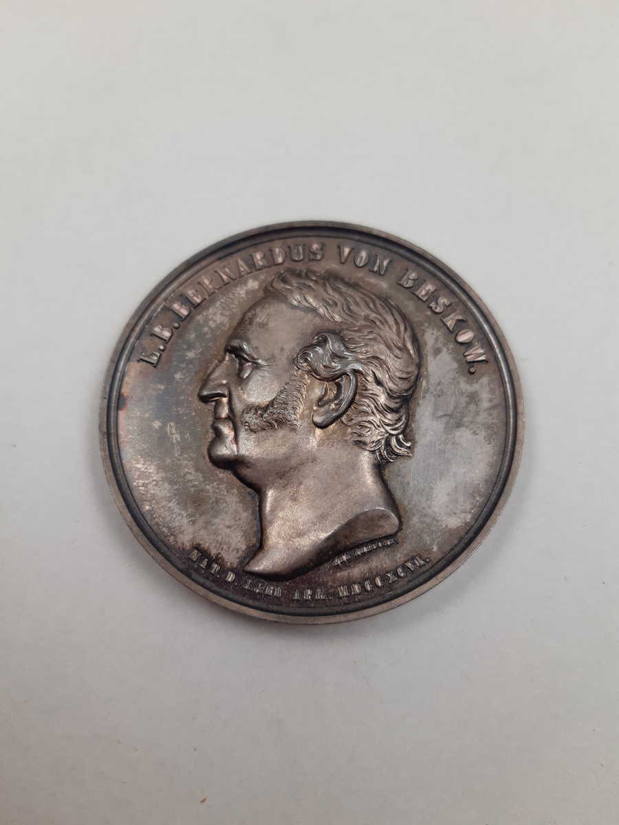 Silvermedalj med reliefdekor, på åtsidan ett mansporträtt i profil och på frånsidan en äldre man med långt skägg sittandes på en tron, han har en harpa vid sin sida och framför honom står två unga kvinnor, en av kvinnorna håller en segerkrans över hans huvud och den andra kvinnan har en penna och en pergamentrulle i sin hand.
