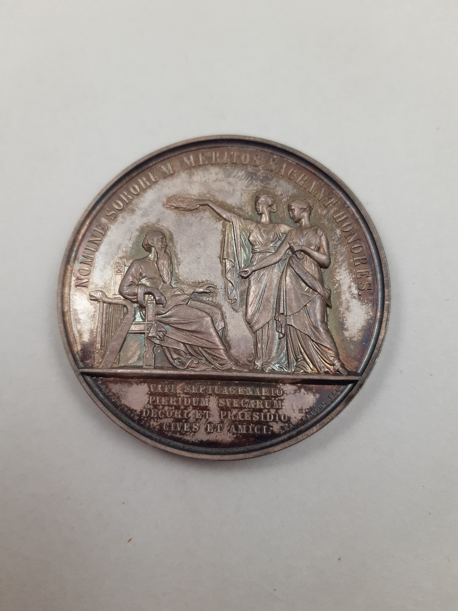 Silvermedalj med reliefdekor, på åtsidan ett mansporträtt i profil och på frånsidan en äldre man med långt skägg sittandes på en tron, han har en harpa vid sin sida och framför honom står två unga kvinnor, en av kvinnorna håller en segerkrans över hans huvud och den andra kvinnan har en penna och en pergamentrulle i sin hand.
