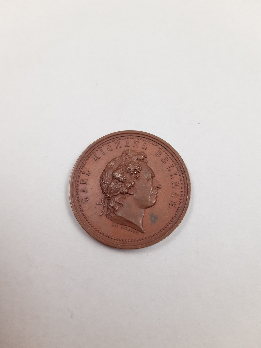 En bronsmedalj, på åtsidan finns ett mansporträtt i profil, frånsidan finns det relief föreställande en halscittra.
