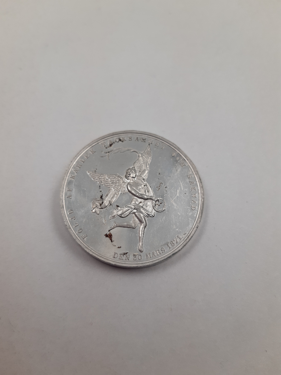 Silvermedalj med reliefdekor, på åtsidan ett drottningporträtt i profil och på frånsidan en ängel som dansar, i hans händer håller han en orm som biter sig själv i svansen och ett palmblad.
