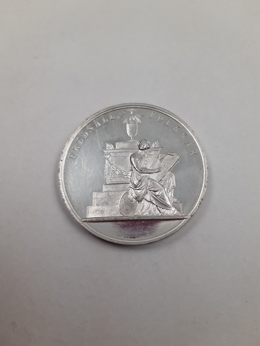 Silvermedalj med reliefdekor, på åtsidan kungaporträtt i profil och på frånsidan en kvinna som sitter på trappan till ett minnesmärke och skriver på en vaxtavla, vid hennes fötter ligger Sveriges statsvapen.
