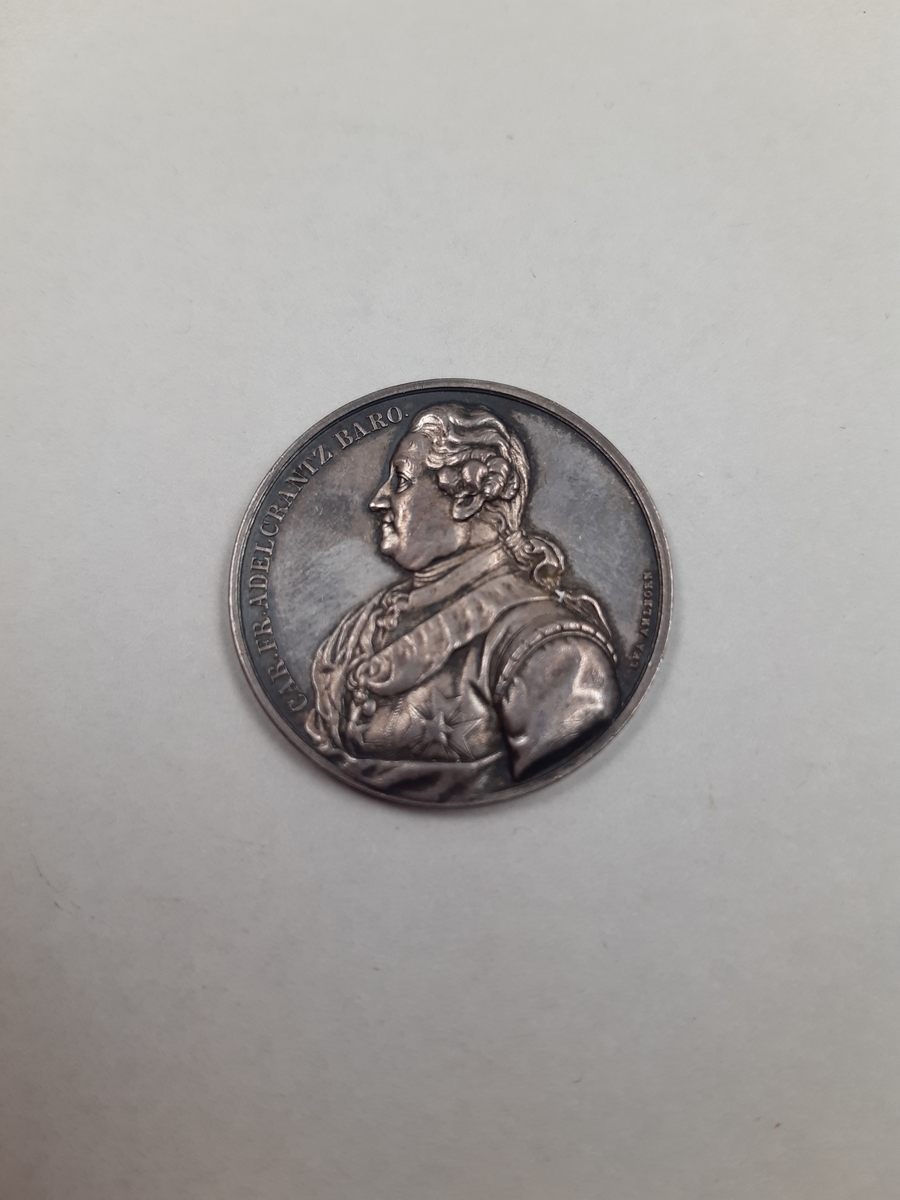 Silvermedalj med reliefdekor, på åtsidan ett mansporträtt i profil och på frånsidan Operahuset i Stockholm.
