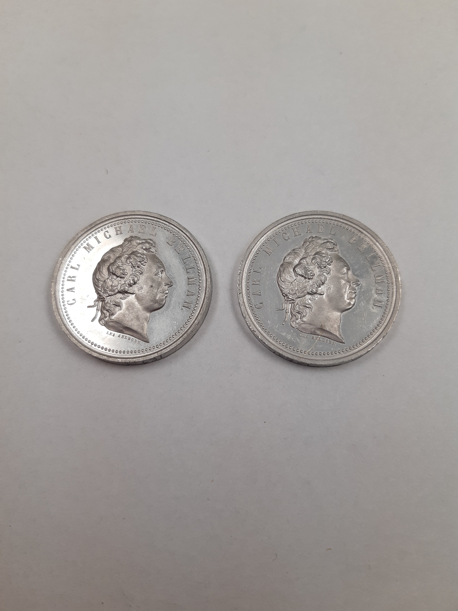 Två silvermedaljer, på åtsidan finns ett mansporträtt i profil, frånsidan finns det relief föreställande en halscittra.
