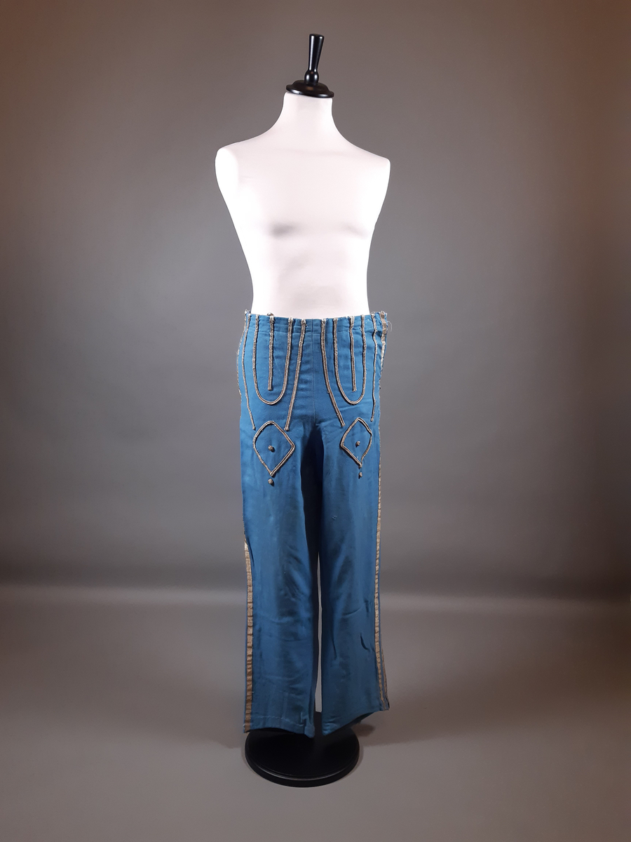 Uniformsbyxor av ull i ljusblå färg. Dekor framtill  med snoddar, snörakeri i guldfärg med en textil kärna. Revärer i guldfärg längs båda byxbenen. Fickor i båda sidor, dragkedja i vänster sida och knappar på insidan runt midjelinningen.
