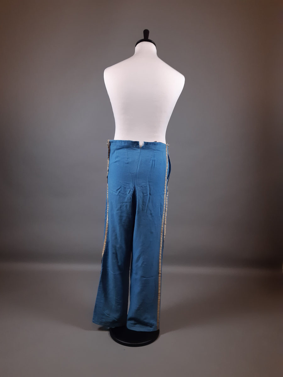 Uniformsbyxor av ull i ljusblå färg. Dekor framtill  med snoddar, snörakeri i guldfärg med en textil kärna. Revärer i guldfärg längs båda byxbenen. Fickor i båda sidor, dragkedja i vänster sida och knappar på insidan runt midjelinningen.
