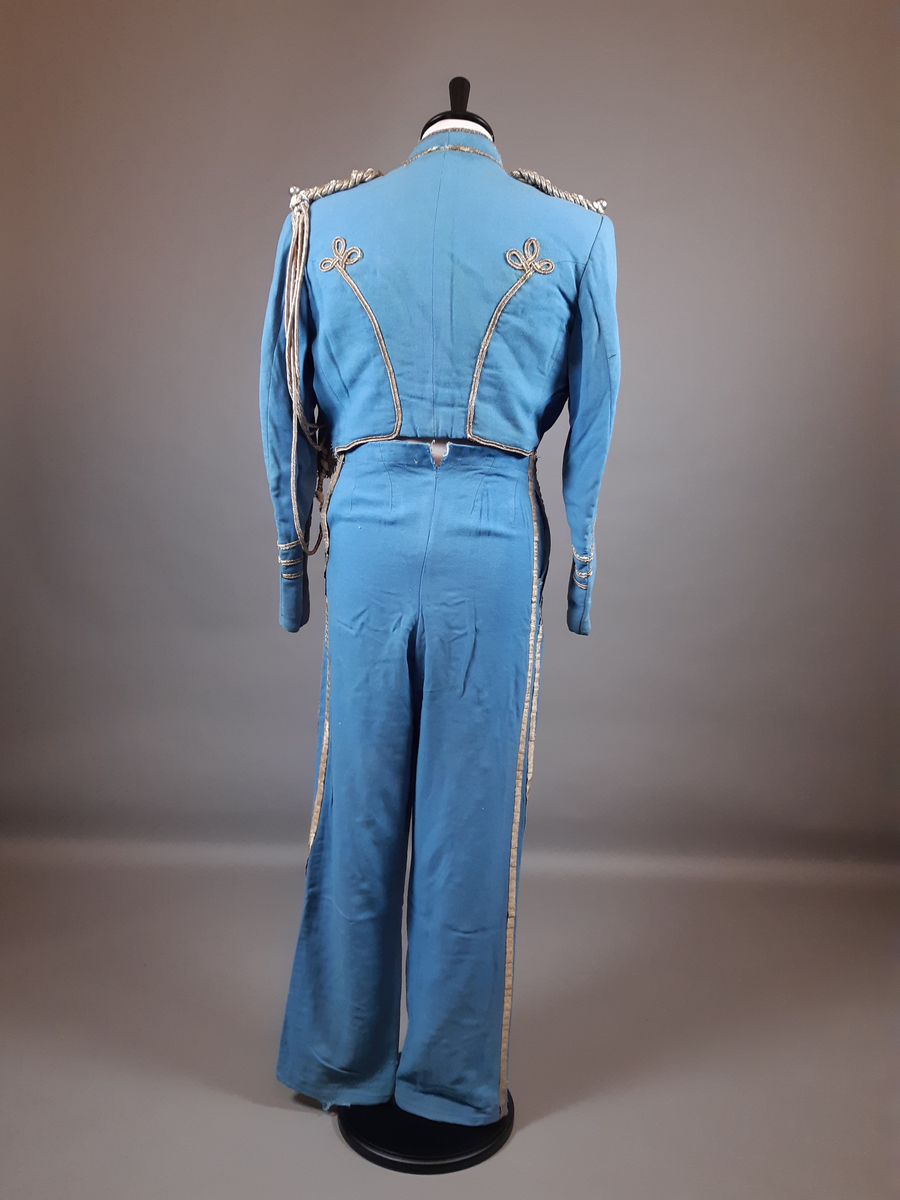 Uniformsjacka av ull i ljusblå färg. Jackan är en kort modell, sk. dolma eller attila. Rikt dekorerad med snoddar, snörmakeri och fransar i guldfärg med en textil kärna. Flätade snoddar på axlarna och en ägiljett runt vänster axel. Klotformade knappar av metall mitt fram, på framstycket och på axlarna. Ståkrage med hyska och hake mitt fram. Fodrad med ett ljusblått tyg med bröstfickor på varje sida. Hakar längs nederkantens insida.
