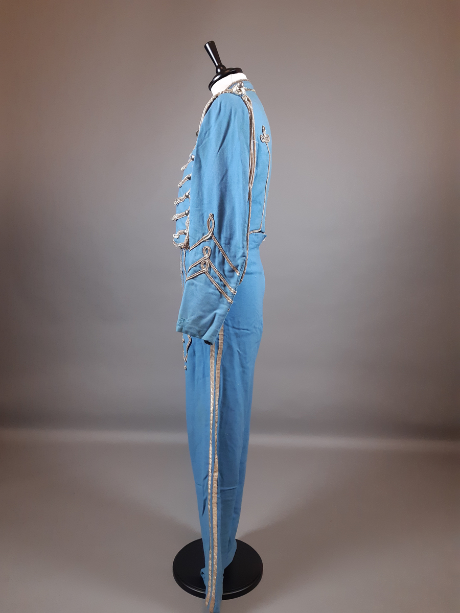 Uniformsjacka av ull i ljusblå färg. Jackan är en kort modell, sk. dolma eller attila. Rikt dekorerad med snoddar, snörmakeri och fransar i guldfärg med en textil kärna. Flätade snoddar på axlarna och en ägiljett runt vänster axel. Klotformade knappar av metall mitt fram, på framstycket och på axlarna. Ståkrage med hyska och hake mitt fram. Fodrad med ett ljusblått tyg med bröstfickor på varje sida. Hakar längs nederkantens insida.
