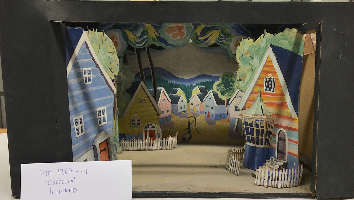 På båda sidor om prosceniet ses två husgavlar. Den vänstra med tre fönster och en dörr. Den högra i blått och orange med en sexkantig veranda, en trappa med staket runtom vilken leder till en ytterdörr på en husfasad. I mellanplanet stiliserade träd med blommor, till vänster ytterligare en husgavel i gult med staket. Fonden är ett torg med kringliggande hus och en vattenpump, bortom detta blånande berg i mjukt vågig stil. 
