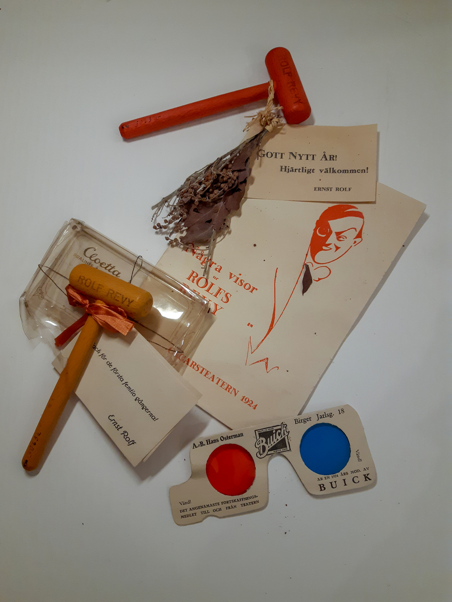 A - Liten röd träklubba med tryck, en torkad liten blomsterbukett och ett visitkort fästade med papperssnöre på ett vishäfte. På vishäftet finns ett stilerat porträtt av Ernst Rolf med konstnärssignatur "E-N" Einar Nerman.
B - Lite ofärgad träklubba med text och ett visitkort fästade med rött sidenband på ett plastetui/hölje för choklad.
C - 3D-glasögon i vit papp med ett rött och ett blått öga i plast. Reklamtext för Buick.
