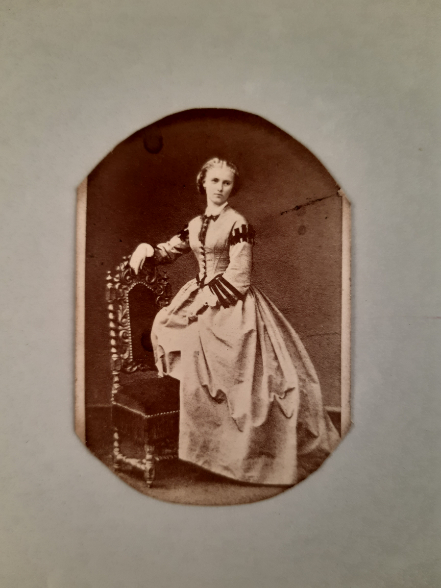 Sepiafärgat fotografi föreställande en kvinna i ljus klänning och ljust uppsatt hår. Hon vilar höger arm på det höga stolsryggen på stolen som står till höger om kvinnan.
