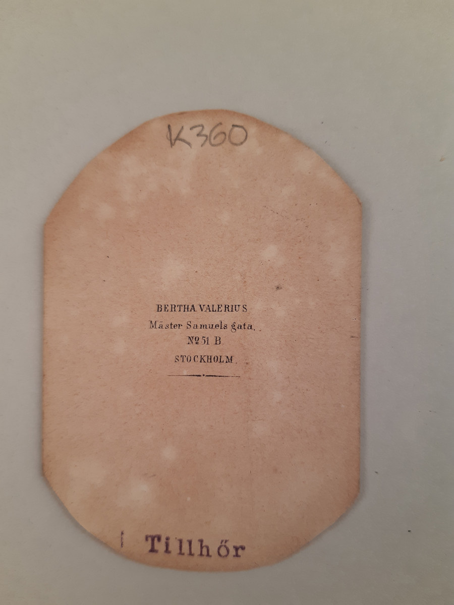 Sepiafärgat fotografi föreställande en kvinna i ljus klänning och ljust uppsatt hår. Hon vilar höger arm på det höga stolsryggen på stolen som står till höger om kvinnan.
