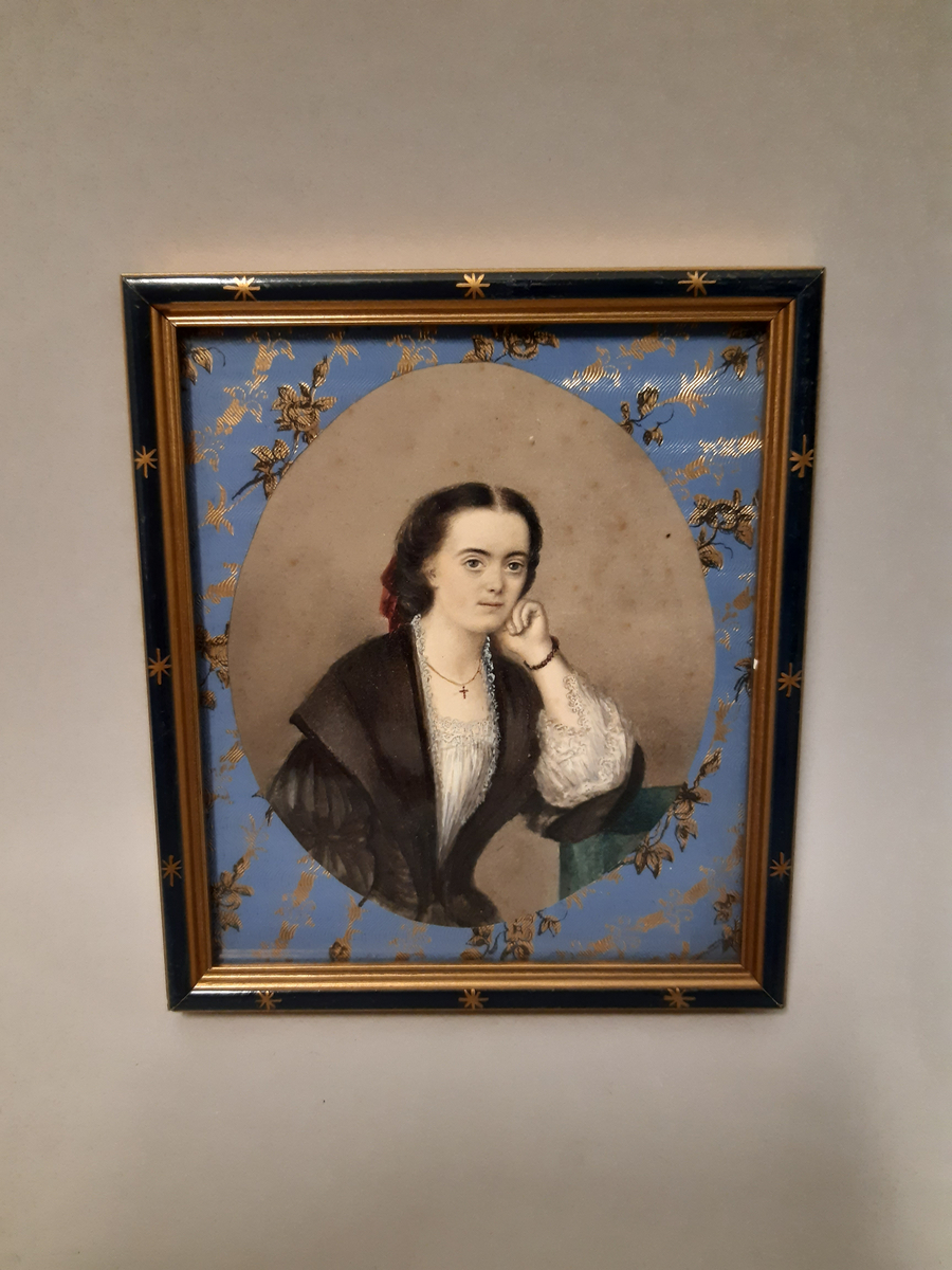 Gouachemålning föreställande en sittande kvinna i mörkt uppsatt hår. Hon är klädd i vit långärmad blus med spets vid ärm- och halslinning. Över blusen bär hon en mörk grönsvart klänning med sjalkrage med vit spets. I håret har hon en mörkröd rosett. Kvinnan vilar huvudet i vänster hand vars armbåge hon stödjer mot ett bord. Runt halsen hänger en tunn kedja med ett litet kors och hon har ett armband gjord av mörkar pärlor kring vänster handled. Passepartout gjord av ljusblått presentpapper med guldtryck. Tavlan har en svart träram med målade dekorationer i guldfärg.
