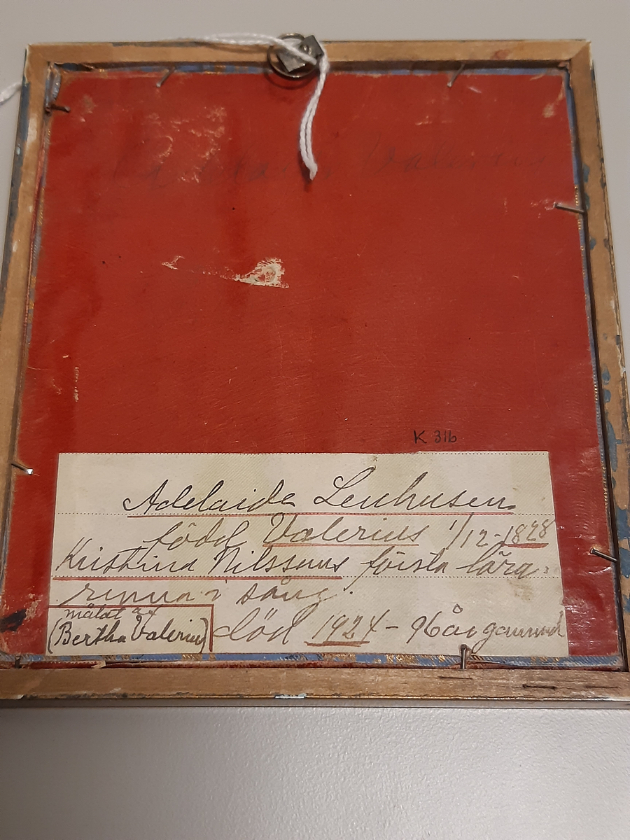 Gouachemålning föreställande en sittande kvinna i mörkt uppsatt hår. Hon är klädd i vit långärmad blus med spets vid ärm- och halslinning. Över blusen bär hon en mörk grönsvart klänning med sjalkrage med vit spets. I håret har hon en mörkröd rosett. Kvinnan vilar huvudet i vänster hand vars armbåge hon stödjer mot ett bord. Runt halsen hänger en tunn kedja med ett litet kors och hon har ett armband gjord av mörkar pärlor kring vänster handled. Passepartout gjord av ljusblått presentpapper med guldtryck. Tavlan har en svart träram med målade dekorationer i guldfärg.
