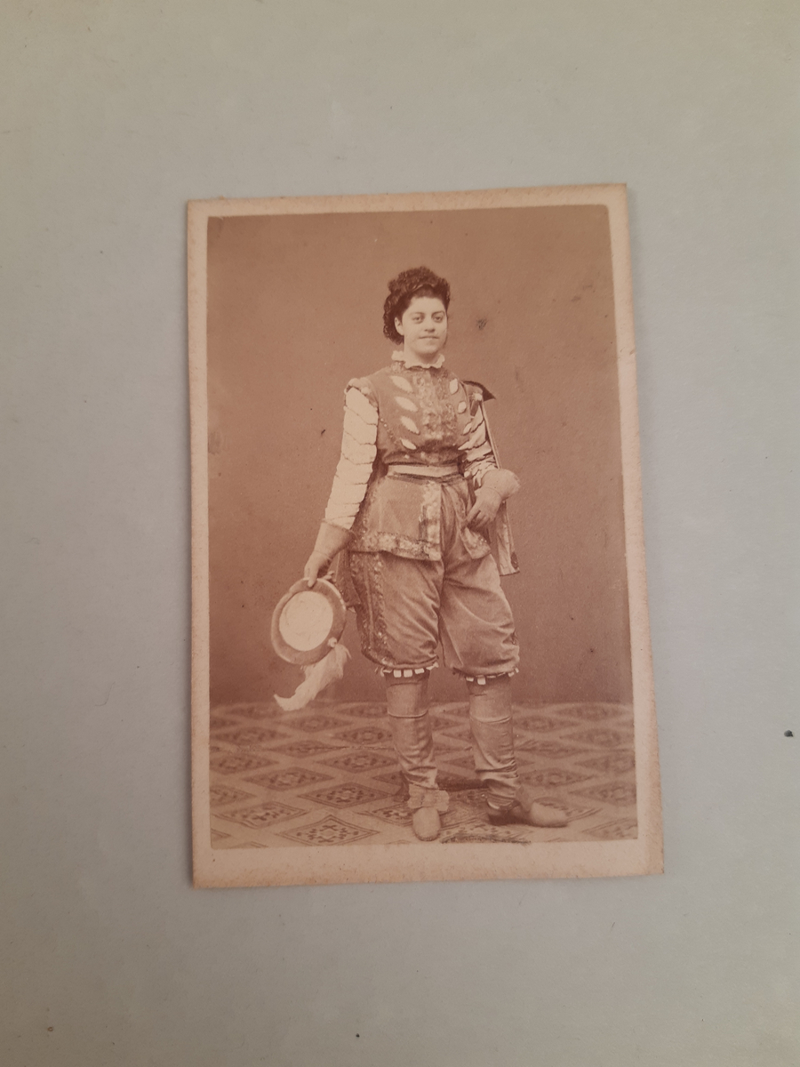 Sepiafärgat fotografi föreställande en kvinna klädd som en renässansyngling i höga stövlar, knäbyxor, väst, jacka och kort mantel. I handen håller hon en fjäderbrydd hatt.
