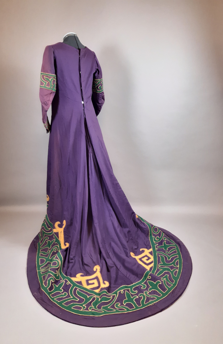 Hellång klänning av kläde i lila färg. Klänningen är längre baktill i ett släp med veckning i ryggen. Dekor av applikationer längs nederkanten, halsringningen och på ärmarna av två ulltyg i grön och gul färg kantad med snoddar i guldfärg. Klänningen är i nationalromantisk stil/jugendstil, tidigt 1900-tal. Fodrad med ett grönt tyg. Knäppning mitt bak med knappar, hyskor, hakar och tryckknappar.
