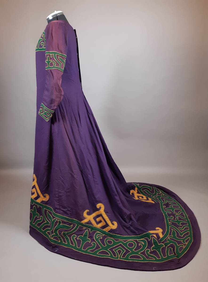 Hellång klänning av kläde i lila färg. Klänningen är längre baktill i ett släp med veckning i ryggen. Dekor av applikationer längs nederkanten, halsringningen och på ärmarna av två ulltyg i grön och gul färg kantad med snoddar i guldfärg. Klänningen är i nationalromantisk stil/jugendstil, tidigt 1900-tal. Fodrad med ett grönt tyg. Knäppning mitt bak med knappar, hyskor, hakar och tryckknappar.
