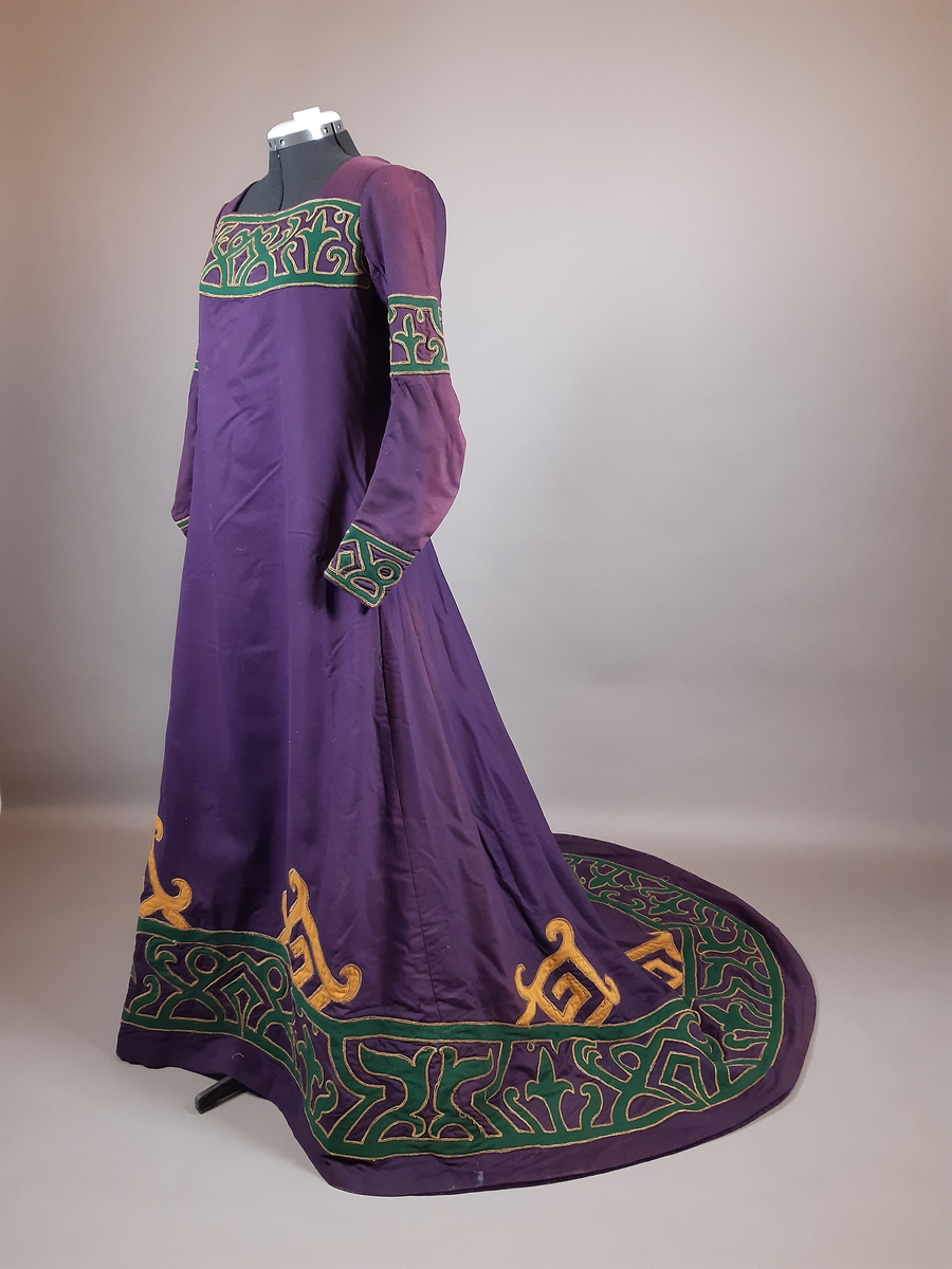 Hellång klänning av kläde i lila färg. Klänningen är längre baktill i ett släp med veckning i ryggen. Dekor av applikationer längs nederkanten, halsringningen och på ärmarna av två ulltyg i grön och gul färg kantad med snoddar i guldfärg. Klänningen är i nationalromantisk stil/jugendstil, tidigt 1900-tal. Fodrad med ett grönt tyg. Knäppning mitt bak med knappar, hyskor, hakar och tryckknappar.
