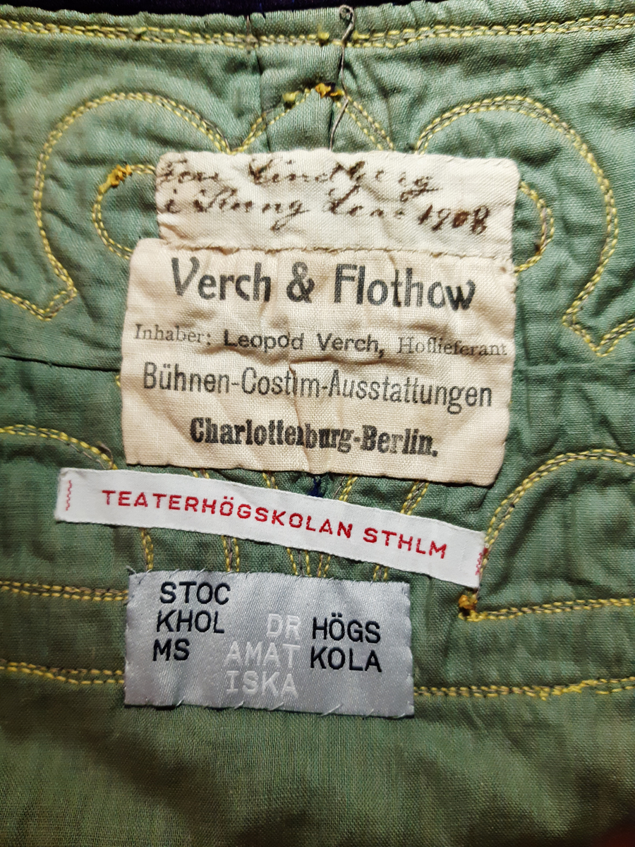 Hellång klänning av kläde i lila färg. Klänningen är längre baktill i ett släp med veckning i ryggen. Dekor av applikationer längs nederkanten, halsringningen och på ärmarna av två ulltyg i grön och gul färg kantad med snoddar i guldfärg. Klänningen är i nationalromantisk stil/jugendstil, tidigt 1900-tal. Fodrad med ett grönt tyg. Knäppning mitt bak med knappar, hyskor, hakar och tryckknappar.
