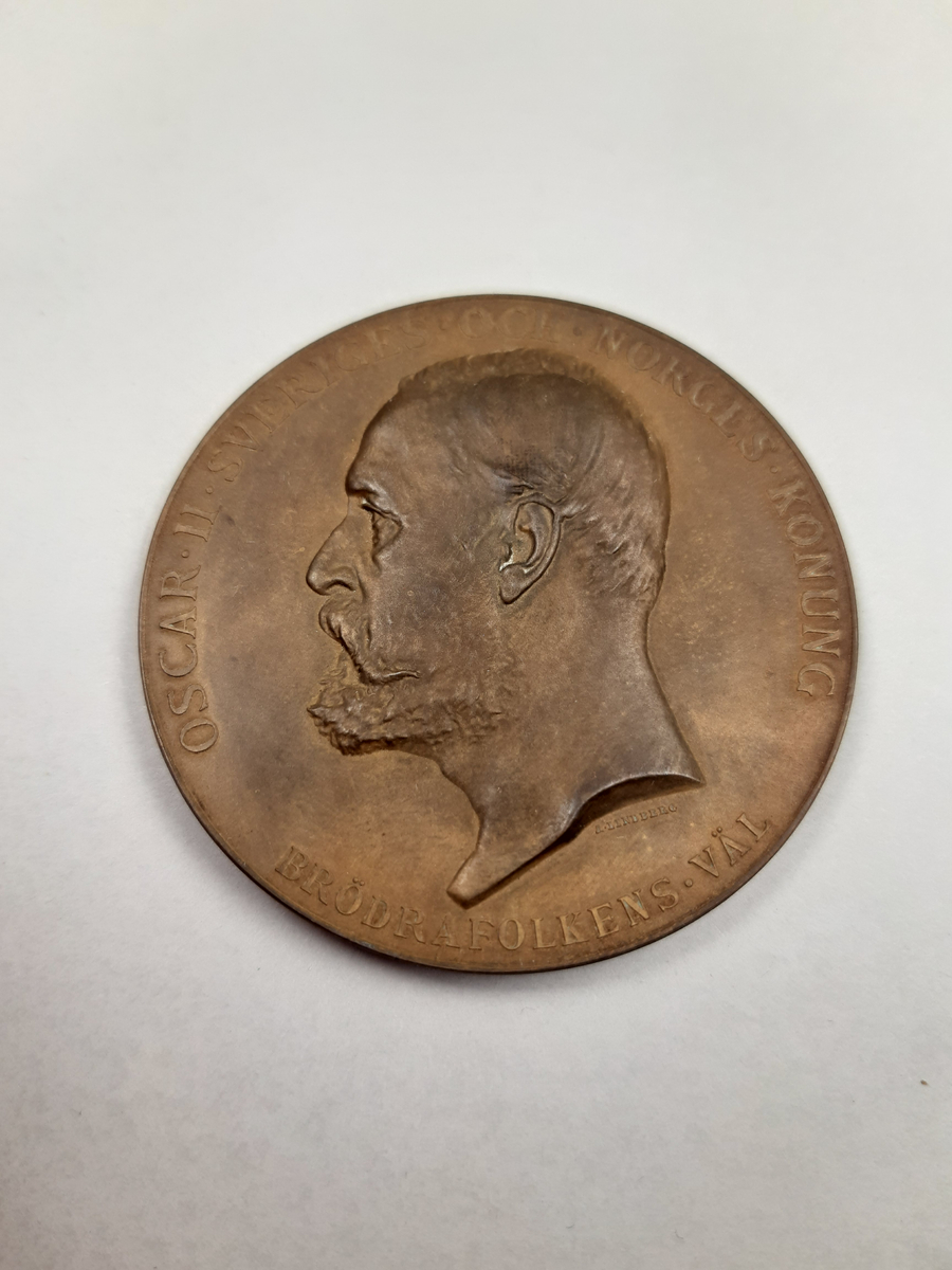 Bronsmedalj med reliefdekor, på åtsidan ett mansporträtt i profil och på frånsidan en segergudinna sittande bland molnen över ett industrilandskap, gudinnan hållet ett palmblad och en lagerkrans i sina händer.
