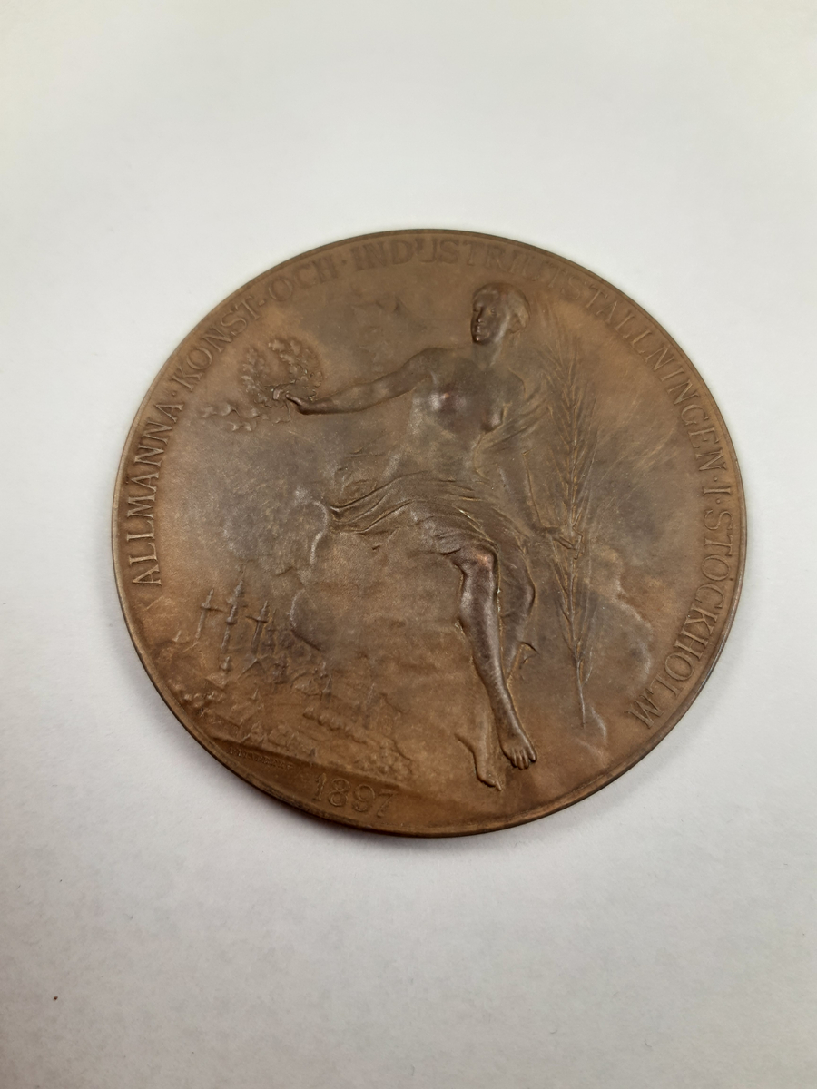 Bronsmedalj med reliefdekor, på åtsidan ett mansporträtt i profil och på frånsidan en segergudinna sittande bland molnen över ett industrilandskap, gudinnan hållet ett palmblad och en lagerkrans i sina händer.
