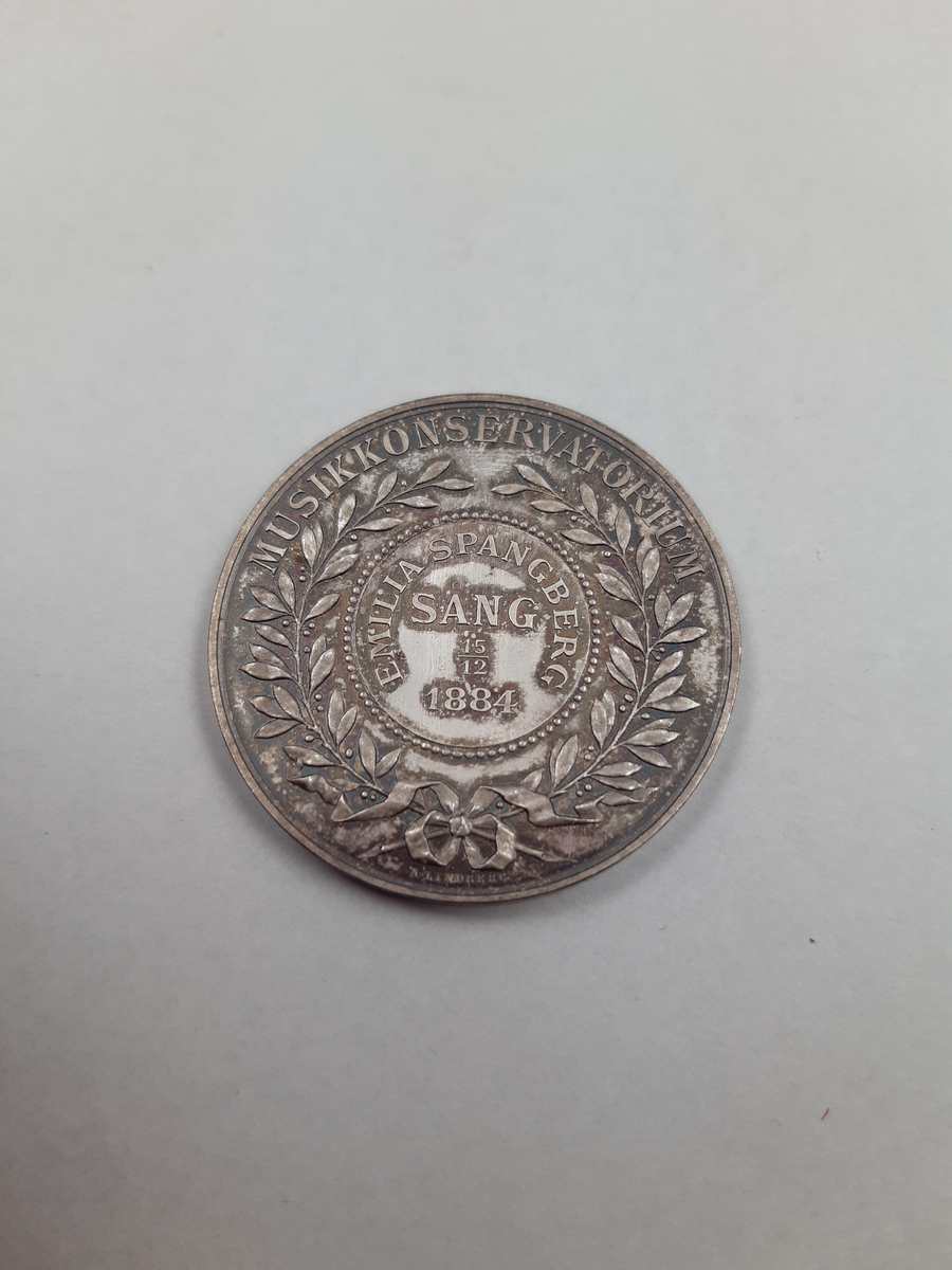 Silvermedalj med reliefdekor, på åtsidan en öppen lagerkrans och på frånsidan en lyra krönt av en strålande stjärna.
