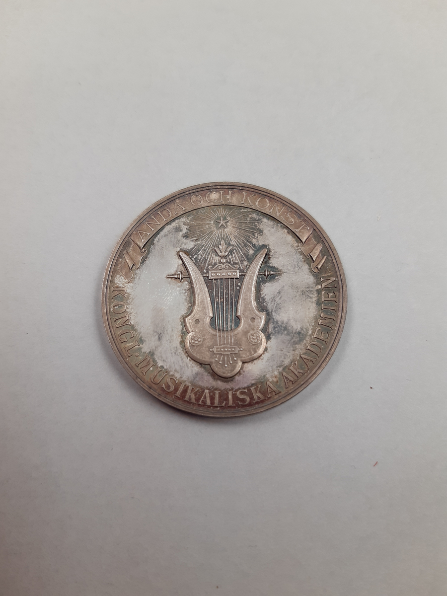 Silvermedalj med reliefdekor, på åtsidan en öppen lagerkrans och på frånsidan en lyra krönt av en strålande stjärna.
