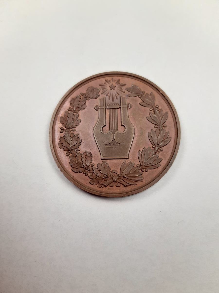 Bronsmedalj med relieddekor, på åtsidan ett mansporträtt i profil och på frånsidan en lyra, en lagerkrans av lagerblad och ekblad samt en strålande stjärna.
