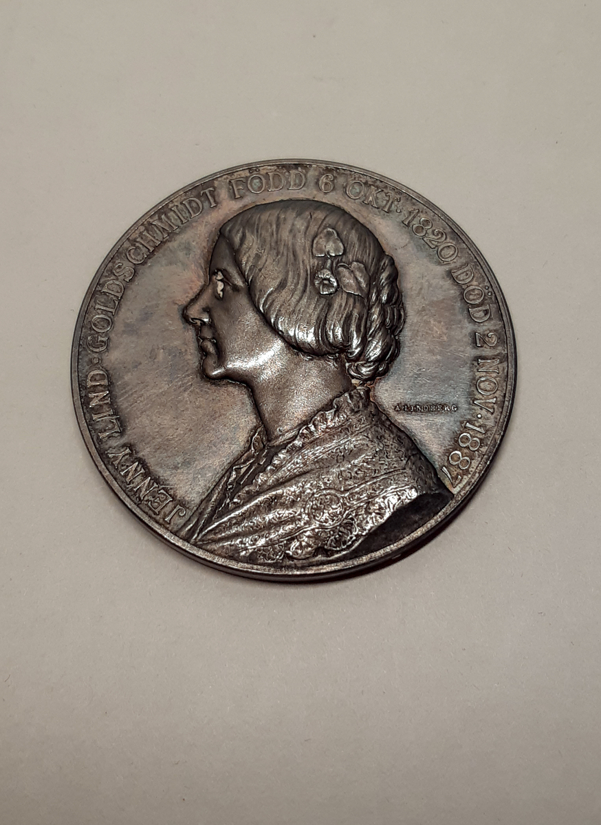 Rund medalj i silver. Motiv på huvudsidan föreställande en äldre kvinna, Jenny Lind,  i profil. Kvinnan har en lindblomma i håtet och en sjal om axlarna. Motiv på baksidan föreställer en sittande ängel skrivandes på en vaxtalva. Ängeln är svept i tyg. Vid hennes fötter ligger en lyra, en trave böcker och en piedestal med en brinnande oljelampa.

