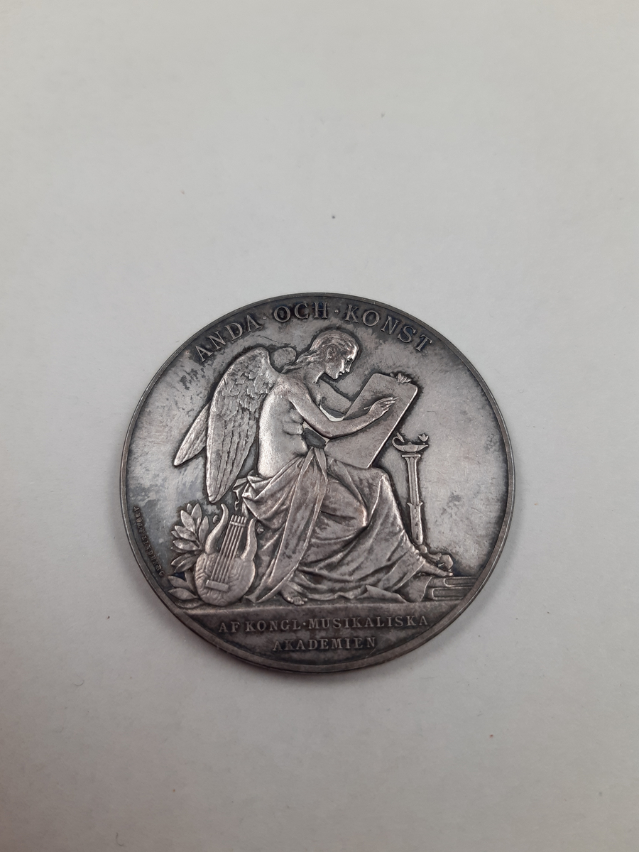 Rund medalj i silver. Motiv på huvudsidan föreställande en äldre kvinna, Jenny Lind,  i profil. Kvinnan har en lindblomma i håtet och en sjal om axlarna. Motiv på baksidan föreställer en sittande ängel skrivandes på en vaxtalva. Ängeln är svept i tyg. Vid hennes fötter ligger en lyra, en trave böcker och en piedestal med en brinnande oljelampa.
