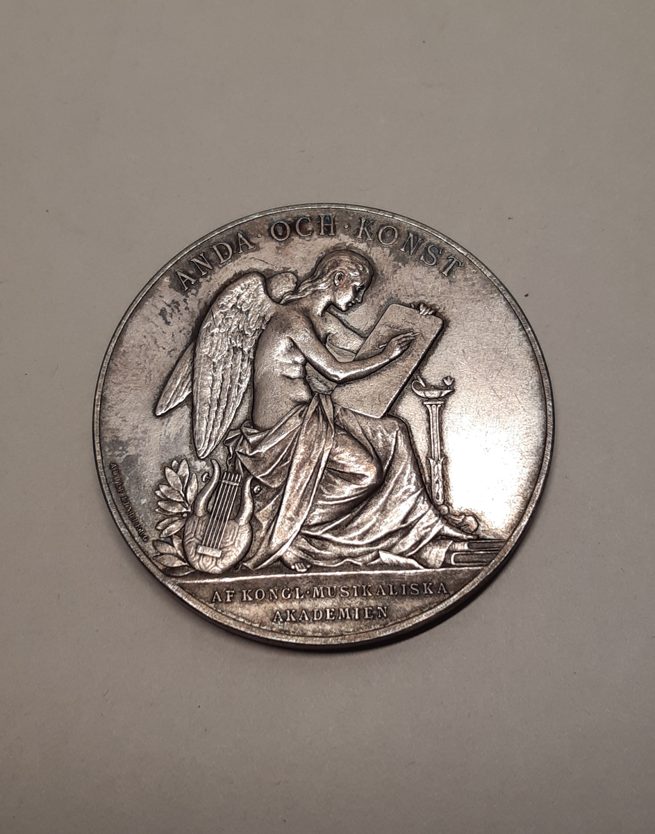 Rund medalj i silver. Motiv på huvudsidan föreställande en äldre kvinna, Jenny Lind,  i profil. Kvinnan har en lindblomma i håtet och en sjal om axlarna. Motiv på baksidan föreställer en sittande ängel skrivandes på en vaxtalva. Ängeln är svept i tyg. Vid hennes fötter ligger en lyra, en trave böcker och en piedestal med en brinnande oljelampa.
