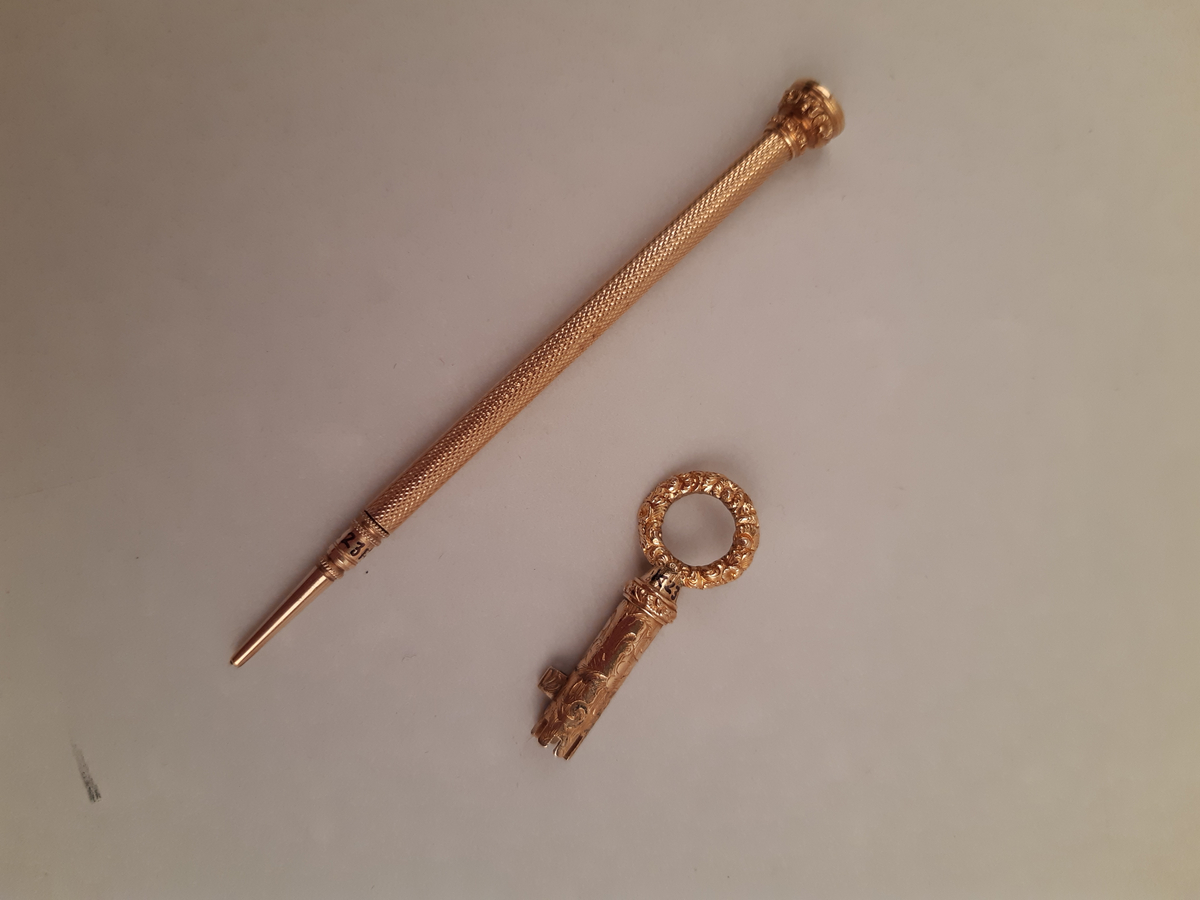 A - Läderklädd bok med lås. Dekorationer i guldtryck på rygg och framsida av pärm.
B - Liten guldnyckel med runt dekorerat handtag.
C - Smal guldpenna med platt guldinfattad elfenbensknopp.
